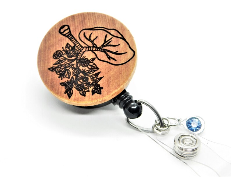 May include: Wooden badge reel with a black and white engraved design of a tree with flowers. The reel has a black retractable cord and a silver clip. The reel is attached to a clear plastic strap with a silver metal ring and a blue rhinestone.