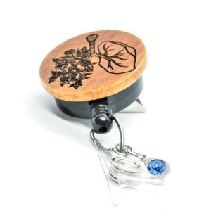 May include: A wooden retractable badge holder with a black retractor and a blue crystal charm. The wooden part features a black and white engraved design of a tree with leaves.