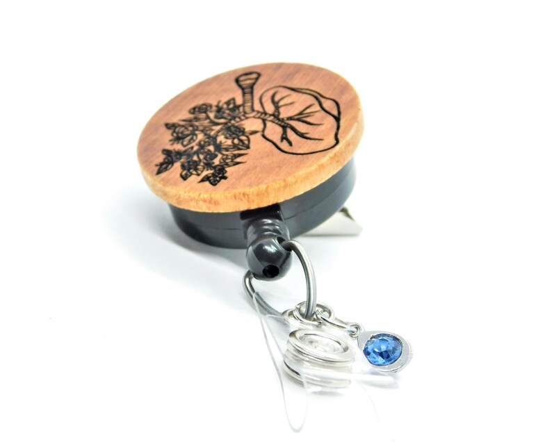 May include: A wooden retractable badge holder with a black retractor and a blue crystal charm. The wooden part features a black and white engraved design of a tree with leaves.