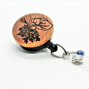 May include: A wooden retractable badge holder with a black engraved floral design and a blue rhinestone charm. The holder is attached to a clear plastic strap.