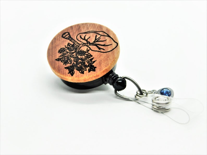 May include: A wooden retractable badge holder with a black engraved floral design and a blue rhinestone charm. The holder is attached to a clear plastic strap.