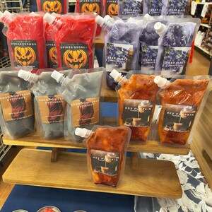 May include: A display of various scented wax melts in pouches and small metal containers. The pouches feature labels with text like "Spiked Eggnog", "Deep Sleep", and "Hey Pumpkin". The melts are in colors like red, purple, orange, and gray.