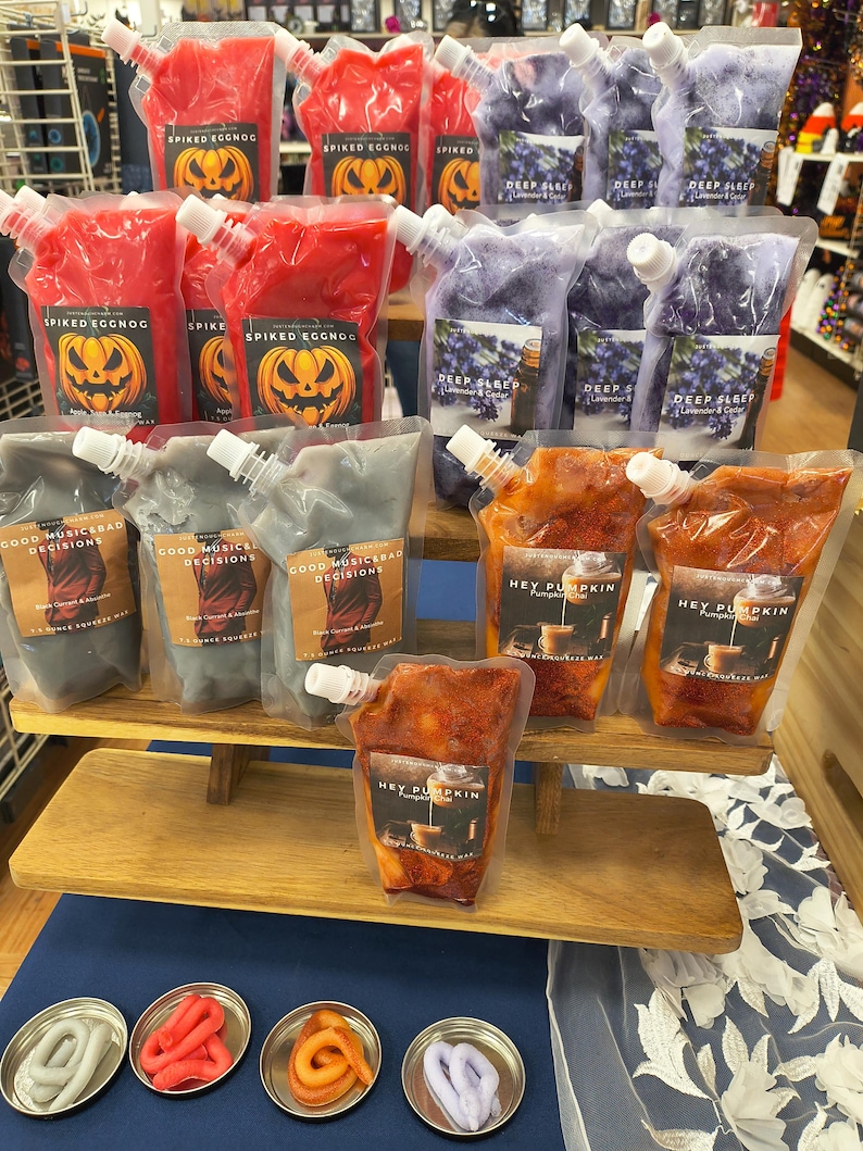 May include: A display of various scented wax melts in pouches and small metal containers. The pouches feature labels with text like "Spiked Eggnog", "Deep Sleep", and "Hey Pumpkin". The melts are in colors like red, purple, orange, and gray.