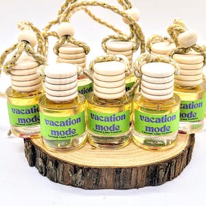 May include: Eight small glass bottles with wooden stoppers and string hangers. Each bottle has a green and white label that reads "vacation mode" with the text "Coconut Lime Verbena" underneath. The bottles are arranged on a wooden slice.