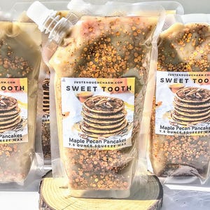 May include: Clear plastic squeeze wax pouches filled with a light brown, speckled substance. Each pouch has a white spout and a label that reads "SWEET TOOTH" and "Maple Pecan Pancakes 7.5 Ounce Squeeze Wax". The pouches are displayed on a wooden slice.