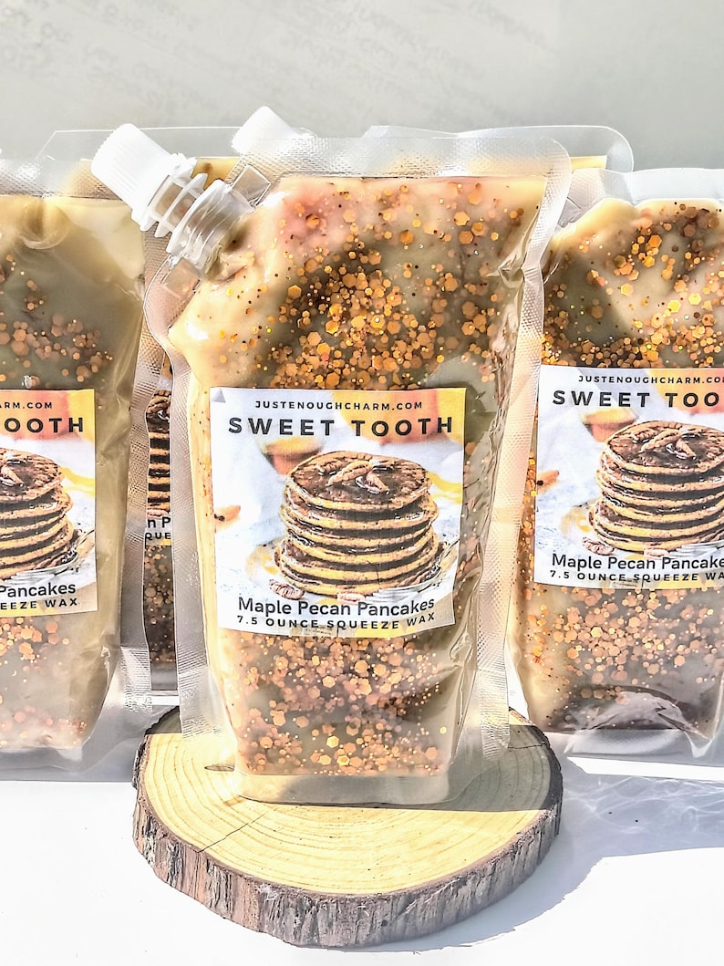 May include: Clear plastic squeeze wax pouches filled with a light brown, speckled substance. Each pouch has a white spout and a label that reads "SWEET TOOTH" and "Maple Pecan Pancakes 7.5 Ounce Squeeze Wax". The pouches are displayed on a wooden slice.