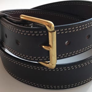Peut inclure: Une ceinture en cuir marron fonc&eacute; avec une boucle dor&eacute;e et des coutures blanches. La ceinture est pli&eacute;e en deux et la boucle est visible.