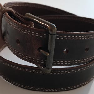 Peut inclure: Une ceinture en cuir marron avec une boucle argent&eacute;e et des coutures blanches. La ceinture est pli&eacute;e en deux et la boucle est visible.
