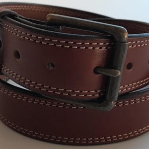 Peut inclure: Une ceinture en cuir marron avec un bord &agrave; double couture et une boucle argent&eacute;e. La ceinture est enroul&eacute;e et la boucle est visible.