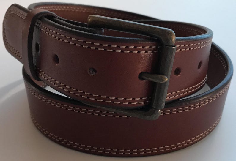 Peut inclure: Une ceinture en cuir marron avec un bord &agrave; double couture et une boucle argent&eacute;e. La ceinture est enroul&eacute;e et la boucle est visible.