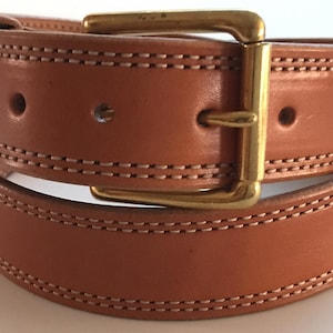 Peut inclure: Une ceinture en cuir marron avec une boucle en laiton et des coutures doubles. La ceinture est pli&eacute;e en deux, montrant les coutures des deux c&ocirc;t&eacute;s.