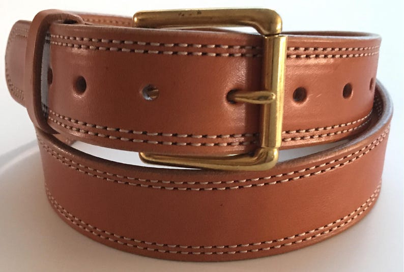 Peut inclure: Une ceinture en cuir marron avec une boucle en laiton et des coutures doubles. La ceinture est pli&eacute;e en deux, montrant les coutures des deux c&ocirc;t&eacute;s.