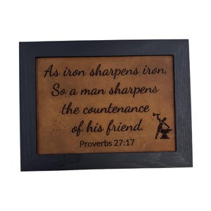 May include: A framed leather plaque with a black frame. The plaque has a brown leather background with the text "As iron sharpens iron, So a man sharpens the countenance of his friend. Proverbs 27:17" engraved in black. The plaque also features a small black silhouette of two people sharpening a piece of metal.