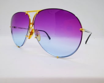 May include: Aviator sunglasses featuring a gold-tone metal frame and a single continuous lens with a vibrant purple to light blue gradient. The bridge and temple arms are constructed from thin gold metal, and the lens is marked with Porsche Design.