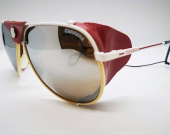 May include: Vintage Carrera sunglasses with silver mirrored lenses, a white frame, and red leather side shields. The sunglasses have a gold rim around the lenses and the Carrera logo is visible on the frame.