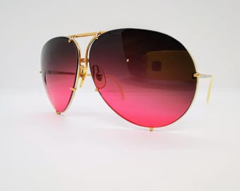 May include: Gold-tone aviator sunglasses with a gradient pink and black lens. The sunglasses have a thin metal frame and a double bridge.