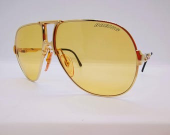 May include: Gold-framed aviator sunglasses with yellow lenses. The word "BOEING" is printed on the top of the frame. The sunglasses have a double bridge and a classic pilot shape, with a light-colored nose bridge and black earpieces.