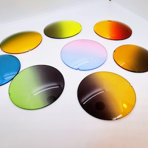 Op de afbeelding: Een assortiment van acht ovale, kleurrijke zonnebrillenglazen. De glazen tonen een verscheidenheid aan kleurenschema's, waaronder geel, blauw, groen, oranje en een roze-blauwe gradi&euml;nt. Elk glas heeft een kleine inkeping aan &eacute;&eacute;n kant.