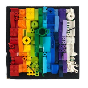 Puede incluir: Un mosaico de colores arcoíris hecho de pequeños bloques de construcción de plástico. Los bloques están dispuestos en un patrón vertical, con cada color representando un tono diferente del arcoíris. El mosaico está colocado sobre un fondo negro.