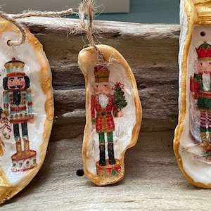 May include: Three oyster shell ornaments painted with nutcracker figures. Each ornament features a different nutcracker design with gold accents and a string for hanging.