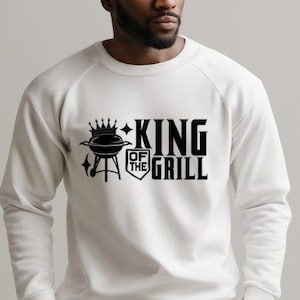 Może przedstawiać: Biała bluza z czarnym nadrukiem graficznym z napisem "King of the Grill" z koroną i grillem.