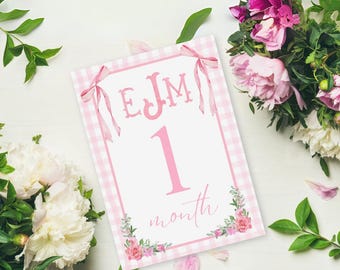 May include: A pink and white gingham-bordered milestone card with the text "EJM 1 month" in pink script. The card is decorated with pink bows and floral accents. The image is surrounded by bouquets of pink and white flowers and green leaves.