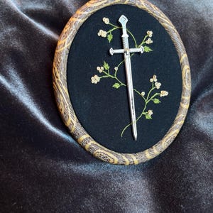 Puede incluir: Un bastidor de bordado ovalado con un fondo de tela negra. Una espada plateada está centrada, con vides verdes bordadas y pequeñas flores blancas. El aro de madera tiene un patrón decorativo.