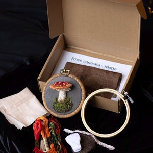 May include: A mushroom embroidery kit with a brown wooden hoop, red and white embroidery floss, a piece of brown felt, a white fabric square, and a wooden embroidery hoop. The kit includes instructions in French.