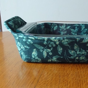 May include: A rectangular glass baking dish with a dark teal fabric cover featuring a leaf pattern. The fabric wraps around the dish, with handles on each end. The dish sits on a wooden surface.