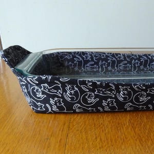 May include: A rectangular glass baking dish inside a black fabric cozy. The fabric is patterned with white cat illustrations. The cozy has handles on each end for easy transport. The dish is on a wooden surface.