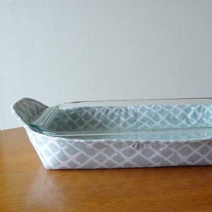 May include: A rectangular glass baking dish inside a fabric carrier. The carrier is light blue and white with a diamond pattern. The handles are curved and padded. The dish is on a wooden surface.