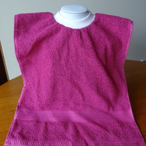 May include: A magenta terry cloth bib, designed to cover the shoulders and chest. The bib has a white, ribbed neckline and a textured surface. The bottom edge features a decorative, horizontal band.