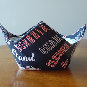 May include: A black fabric bowl with red and white lettering that says "Cleveland Guardians" and "Grand". The bowl is designed to hold a hot beverage mug or bowl.