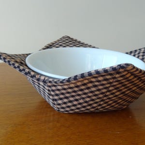 May include: A white ceramic bowl sits inside a black and tan gingham fabric bowl cozy. The cozy has four pointed corners. The bowl and cozy are on a wooden surface.