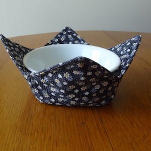 May include: A white ceramic bowl sits inside a navy blue fabric bowl cozy. The cozy is decorated with a pattern of small white, yellow, and gray flowers. The cozy is designed to hold the bowl and protect the user's hands from heat.