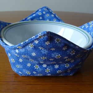 May include: A blue fabric bowl cozy with a white daisy print. The cozy is designed to hold a bowl and keep it warm.