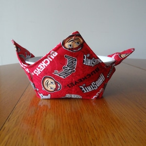 May include: A red fabric bowl cozy featuring a repeating pattern of the Ohio State Buckeyes logo and the words "OHIO STATE" and "BUCKEYES" in white. The cozy is designed to hold a bowl and has pointed flaps.