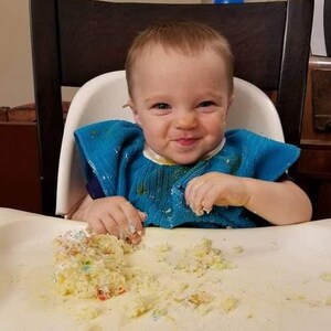 Toddler boy in high chair eating cake and wearing a pullover towel bib.