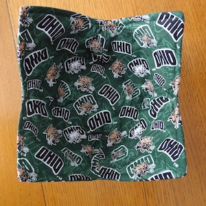May include: Green fabric bowl cozy with a repeating pattern of the word "OHIO" in white and the Ohio University Bobcats logo in brown and white. The cozy is square-shaped and sits on a wooden surface.