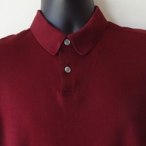 May include: A sleeveless polo shirt in a deep burgundy color. The garment features a classic collar and a front placket with two small, light-colored buttons. The fabric appears smooth and solid, with no visible patterns or additional textures.