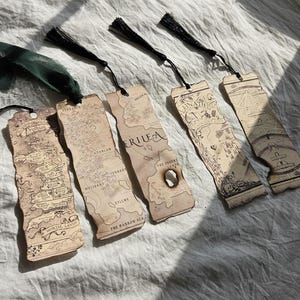 May include: A collection of five vintage-style bookmarks with black tassels. Each bookmark features a detailed map design with text, including "Druthian," "Erilea," and "Lunath." The bookmarks are made of a light brown material.