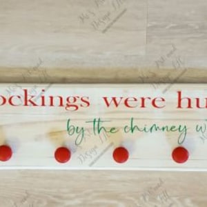 May include: A white wooden sign with the text "the stockings were hung... by the chimney with care" in red and green lettering. The sign has eight red hooks for hanging stockings.
