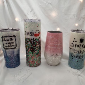 May include: Four different glitter tumblers with various sayings. The tumblers are white with glitter and have different colored gradients. The sayings on the tumblers are "Dance like no one is watching", "I am so freaking merry", "Not all who wander are lost, my words are speaking", and "I'm so freaking cold".
