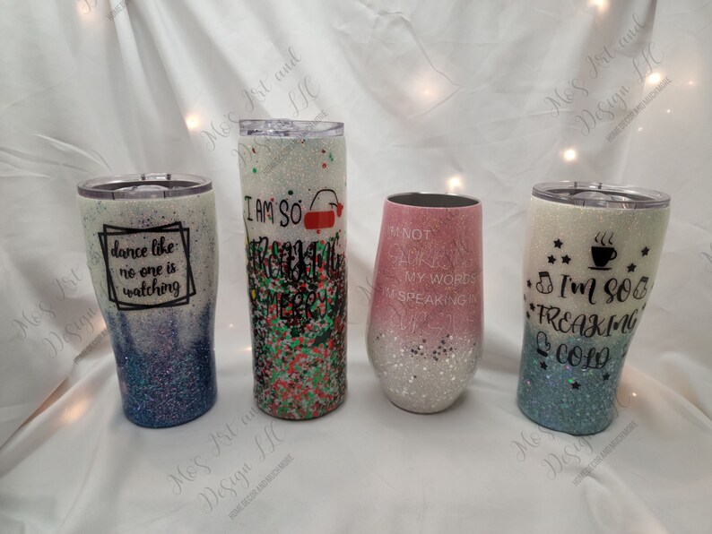 May include: Four different glitter tumblers with various sayings. The tumblers are white with glitter and have different colored gradients. The sayings on the tumblers are "Dance like no one is watching", "I am so freaking merry", "Not all who wander are lost, my words are speaking", and "I'm so freaking cold".