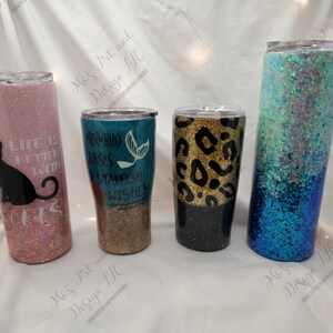 May include: Four different glitter tumblers with various designs. The tumblers are all the same size and shape. The first tumbler is pink with a black cat and the text "Life is better with cats". The second tumbler is teal with a mermaid and the text "Mermaid kisses and starfish wishes". The third tumbler is black with a gold leopard print. The fourth tumbler is blue with a white and blue ombre glitter design.