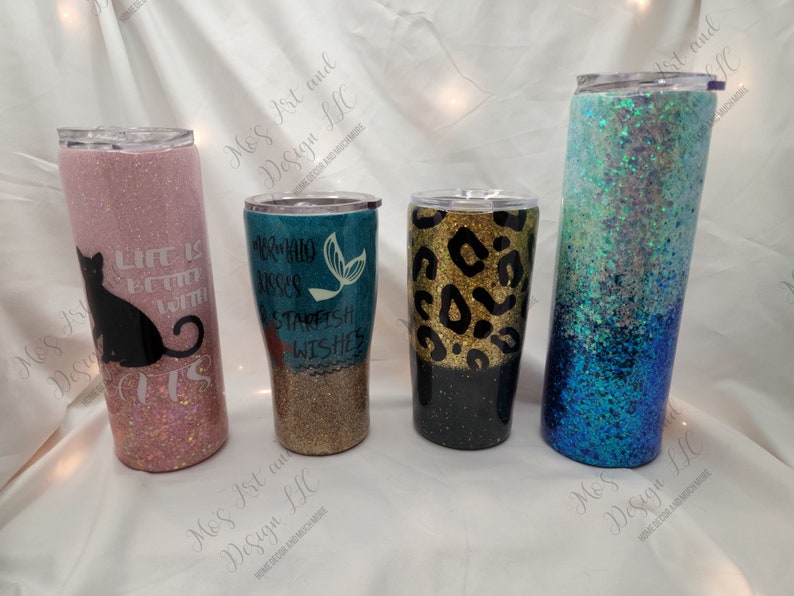 May include: Four different glitter tumblers with various designs. The tumblers are all the same size and shape. The first tumbler is pink with a black cat and the text "Life is better with cats". The second tumbler is teal with a mermaid and the text "Mermaid kisses and starfish wishes". The third tumbler is black with a gold leopard print. The fourth tumbler is blue with a white and blue ombre glitter design.