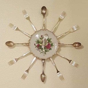 May include: A decorative clock made from a white porcelain plate with a floral design and gold trim. The clock face is surrounded by twelve silver forks and spoons.