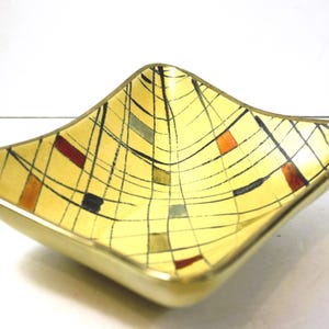 May include: A square ceramic dish with a yellow background and a geometric pattern of black, red, and orange lines. The dish has a gold rim.