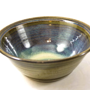 May include: A handcrafted ceramic bowl with a glossy finish. The bowl displays a gradient of colours, including blue, green, and brown. The interior has a smooth, reflective surface, while the exterior has a textured, earthy appearance. Suitable for serving food.