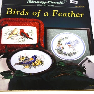 May include: Stoney Creek Collection Book 116, Birds of a Feather cross stitch pattern book. The cover features three framed cross stitch designs of birds. The first design is a red cardinal and a yellow bird on a branch. The second design is a bluebird and a brown bird on a branch. The third design is a blue bird with yellow accents on a branch.